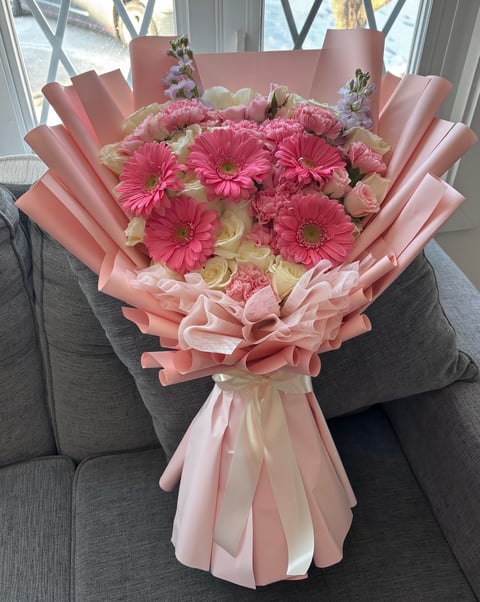 Pink floral bouquet with hot pink gerberas, roses, and carnations wrapped in layers of pale pink tissue paper, displayed on a gray couch by a window