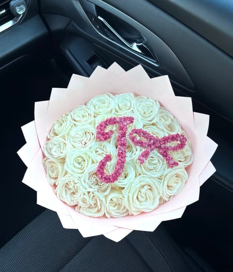 Bouquet of cream-colored roses arranged in a circle with hot pink flowers forming the number 55, wrapped in white paper, displayed on a car seat
