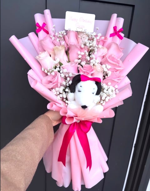Hand holding a pink and white flower bouquet with a plush dog toy, pink ribbons, and babys breath flowers wrapped in pink paper
