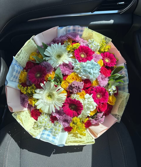 Colorful bouquet of mixed flowers including gerberas, roses, and daisies wrapped in pastel paper sitting in a car seat