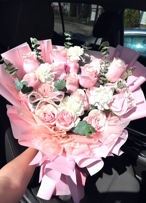 Person holding a large bouquet of pink roses, white carnations, and eucalyptus wrapped in pink paper inside a car