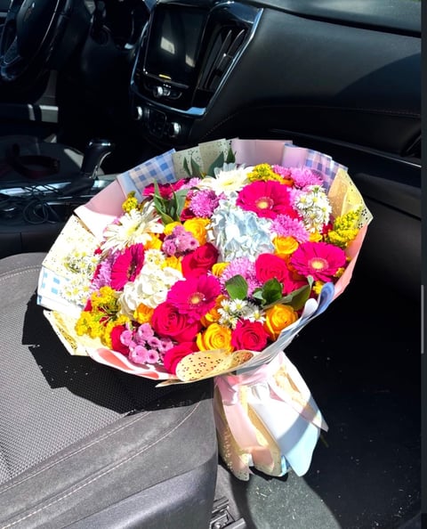 Vibrant bouquet of pink, red, yellow, and white flowers with green foliage wrapped in pink paper, placed on car seat interior